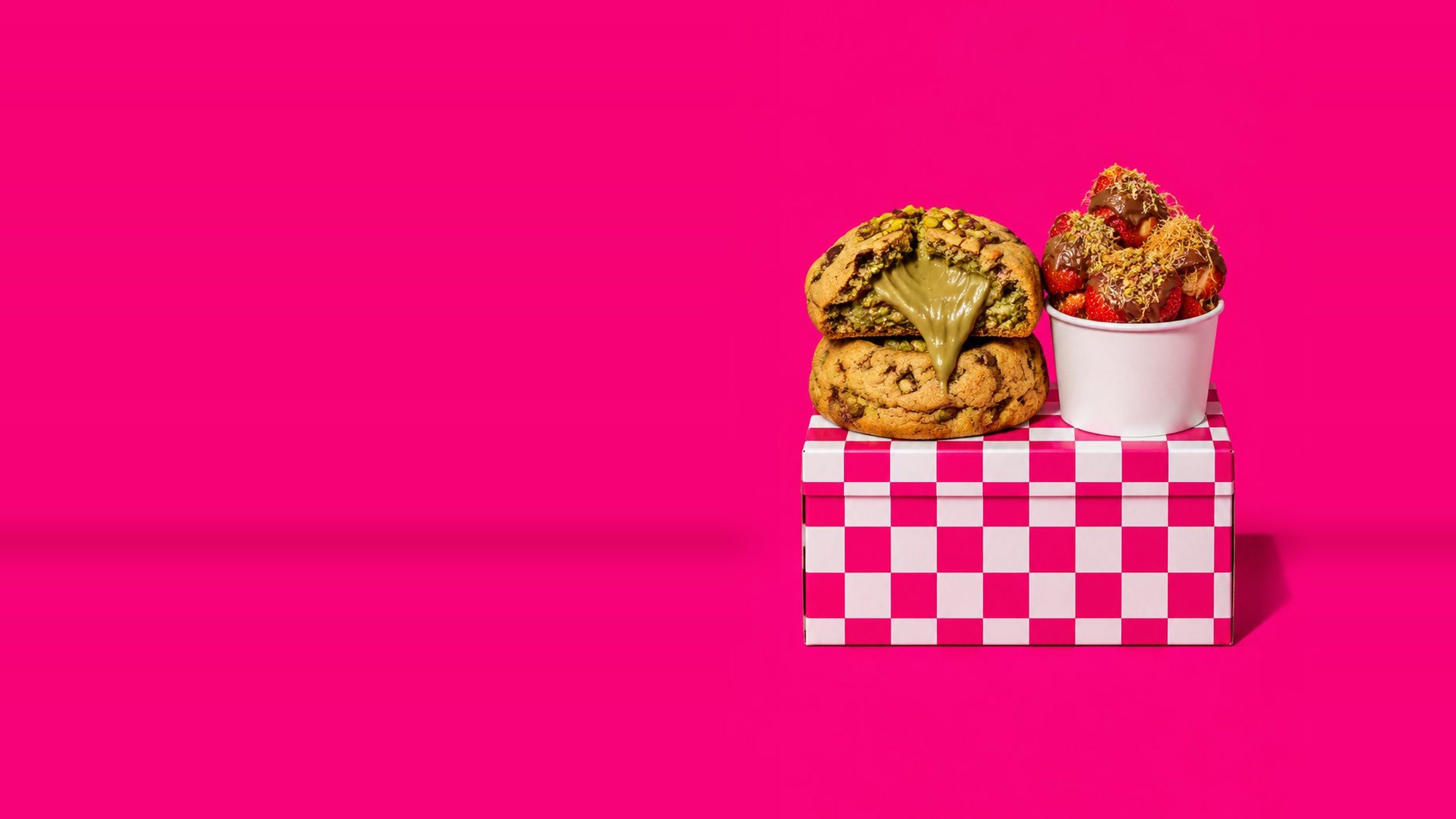 Cookies XXL fourrés à la pistache et pot de fraises au chocolat devant un damier rose et blanc